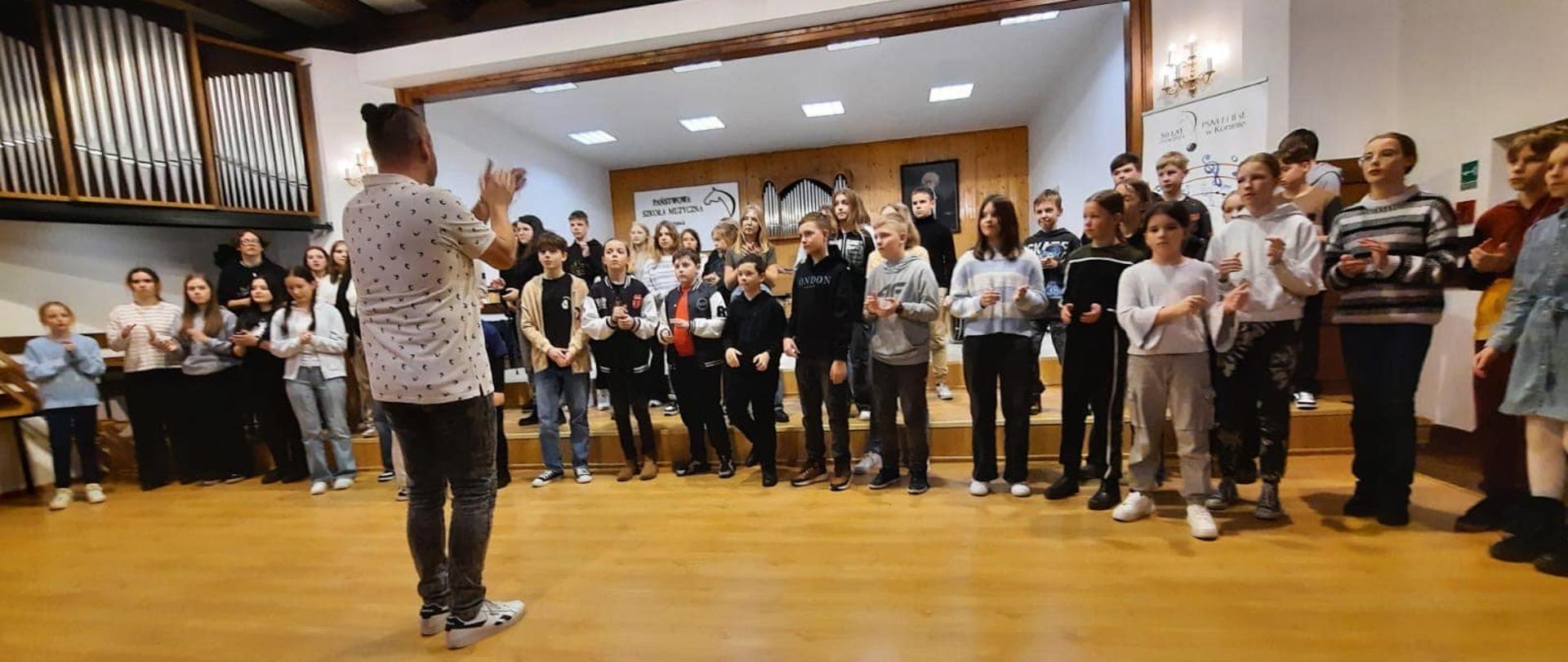 Grupa uczniów stoi na scenie PSM w Koninie przodem do prowadzącego warsztaty, który klaszcze.