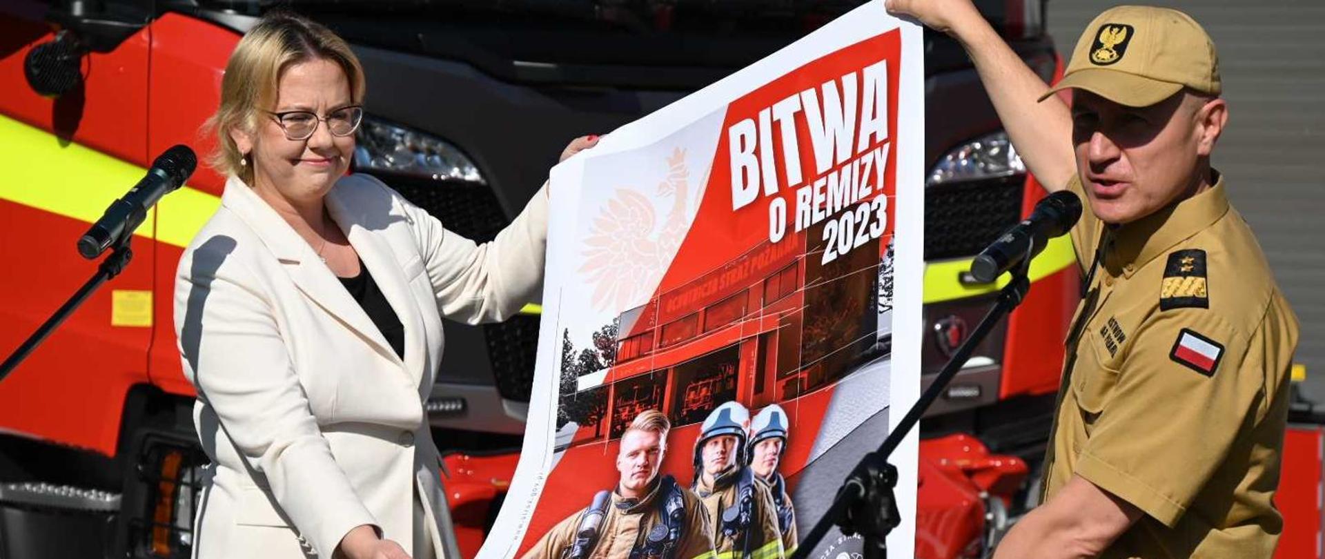 Z lewej Pani Minister Anna Moskwa w białym żakiecie i spodniach, z prawej Komendant Główny PSP w żółtym mundurze służbowym, obydwoje trzymają plakat akcji Bitwa o Remizy. W tle pojazdy pożarnicze ciężarowe koloru czerwonego.