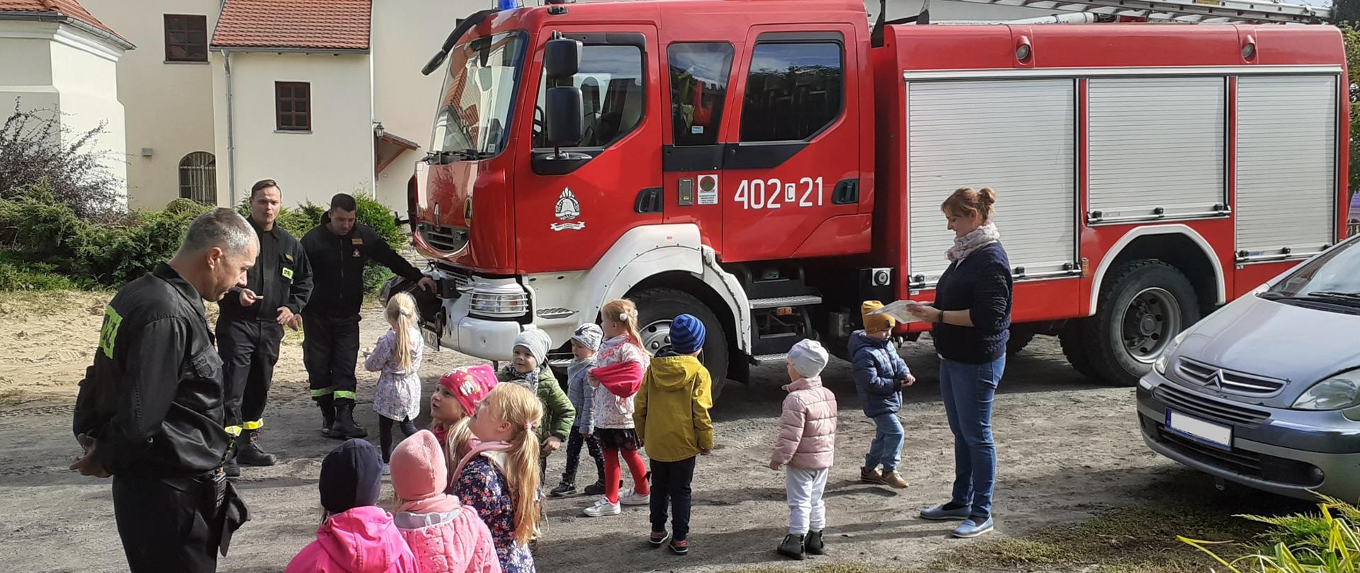 Na zdjęciu widać samochód ratowniczo-gaśniczy oraz grupę przedszkolaków, biorących udział w pokazie sprzętu. Dzieci rozmawiają ze strażakami. W grupie przedszkolaków stoi opiekunka, trzymająca w ręku kartkę z podziękowaniem dla strażaków. Spotkanie odbywa się na parkingu przed budynkiem przedszkola. W tle widać budynek kościoła.