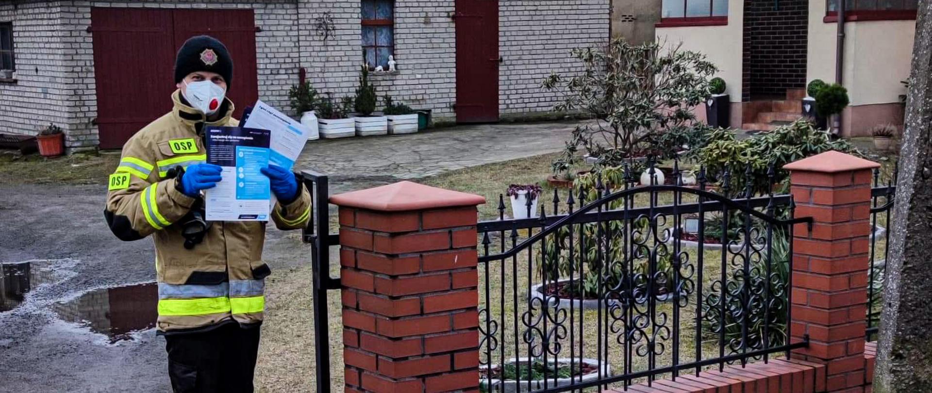 Na zdjęciu znajduje się strażak ubrany w ubranie specjalne, który ma założoną maseczkę ochronną. Strażak stoi przy płocie trzymając w rękach ulotkę informacyjną. W tle znajdują się budynki, a zdjęcie wykonano w porze dziennej przy zachmurzonym niebie. 
