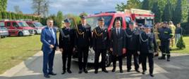 Funkcjonariusze Państwowej Straży Pożarnej, druhowie z Ochotniczej Straży Pożarnej i urzędnicy stoją przed nowym pojazdem pożarniczym. W tle zgormadzeni strażacy i inne pojazdy pożarnicze.