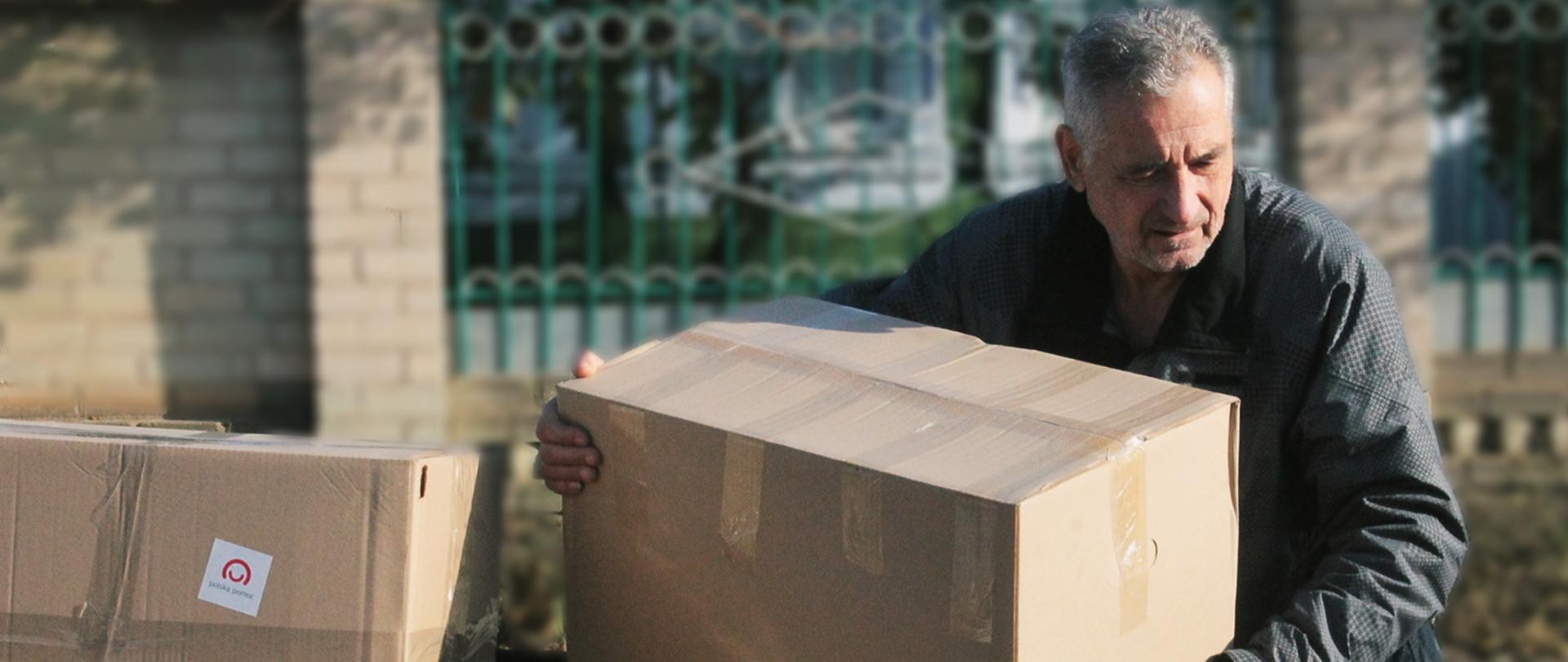 Starszy mężczyzna podnosi jedno z pudeł z pomocą materialną