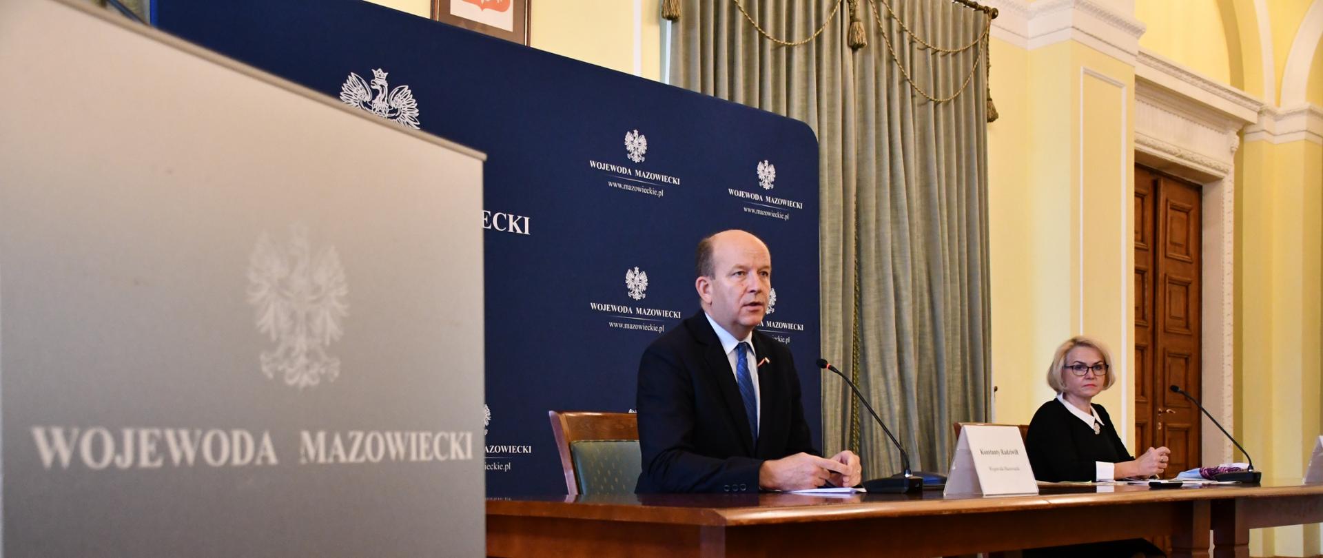Na zdjęciu z lewej strony widoczny jest fragment mównicy z logo Wojewody Mazowieckiego, które przestawia orła w koronie i napis Wojewoda Mazowiecki. Obok widoczni są siedzący przy stole Wojewoda Konstanty Radziwiłł oraz Zastępca Mazowieckiego Państwowego Wojewódzkiego Inspektora Sanitarnego Agata Wolska, który udzielają informacji mediom. Dziennikarze nie są widoczni na zdjęciu.