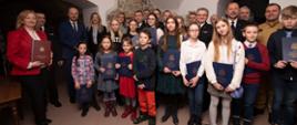 Zdjęcie przedstawia fotografie grupową laureatów konkursu, którzy w dłoniach trzymają nagrody wraz ze strażakami ubranymi w mundury wyjściowe oraz przybyłymi gośćmi.