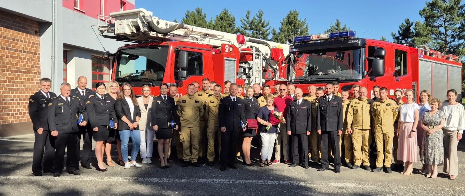 Na zdjęciu znajdują się osoby ustawione do zdjęcia na tle samochodów strażackich ubrane w mundury i ubiory cywilne