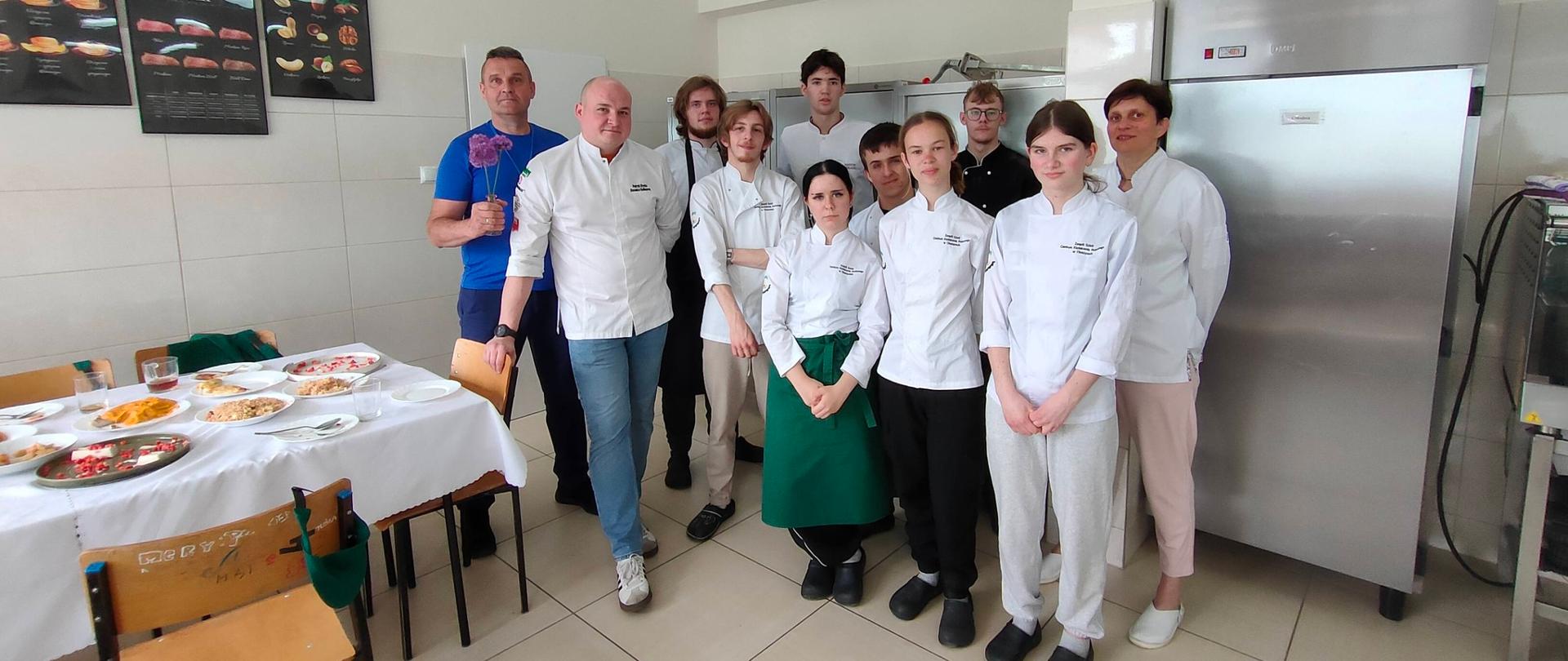 Grupowe zdjęcie uczestników warsztatów kulinarnych Develey w szkolnej pracowni gastronomicznej. Na pierwszym planie stoją uczniowie ubrani w białe fartuchy kucharskie. Z lewej strony stoi mężczyzna w niebieskiej koszulce - pan Adam Socha - przedstawiciel handlowy firmy Develey, obok niego ekspert kulinarny w białym kucharskim kitlu. W tle widoczne są sprzęty kuchenne, lodówka przemysłowa oraz plakaty tematyczne na ścianie. Na stole po lewej stronie widoczne są przygotowane podczas warsztatów potrawy. 19 maja 2026 r.