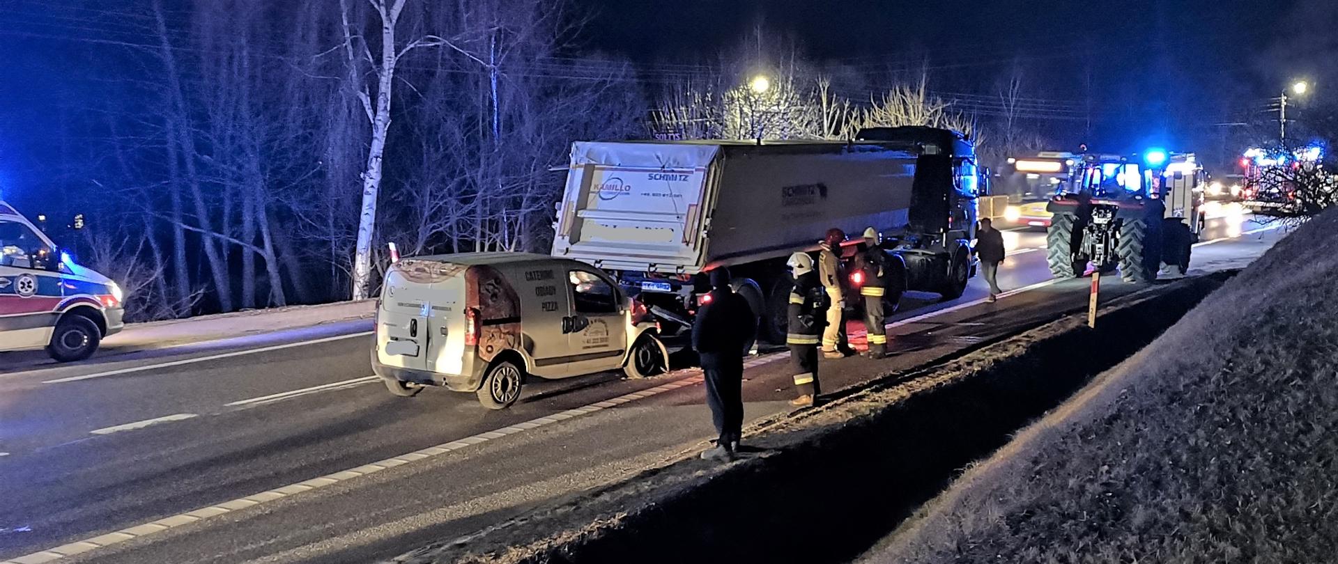 Na zdjęciu przedstawiono drogę krajowa na której doszło do zderzenia dwóch samochodów. Fiat Fiorino znajduje się za naczepą ciągnika siodłowego Scania. Widać zniszczone przednie koło. Na poboczu prawej strony, grupa strażaków i policjant, po drugiej stronie ulicy stoi ambulans Ratownictwa Medycznego.