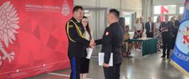 Mazowiecki Komendant Wojewódzki PSP nadbryg. Artur Gonera składa gratulacje bryg. Karolowi Marcikowskiemu po wręczeniu aktu powołania na stanowisko Komendanta Powiatowego PSP w Garwolinie. Strażacy ściskają sobie dłonie na tle okolicznościowej ścianki Państwowej Straży Pożarnej, a w tle znajdują się uczestnicy uroczystości.