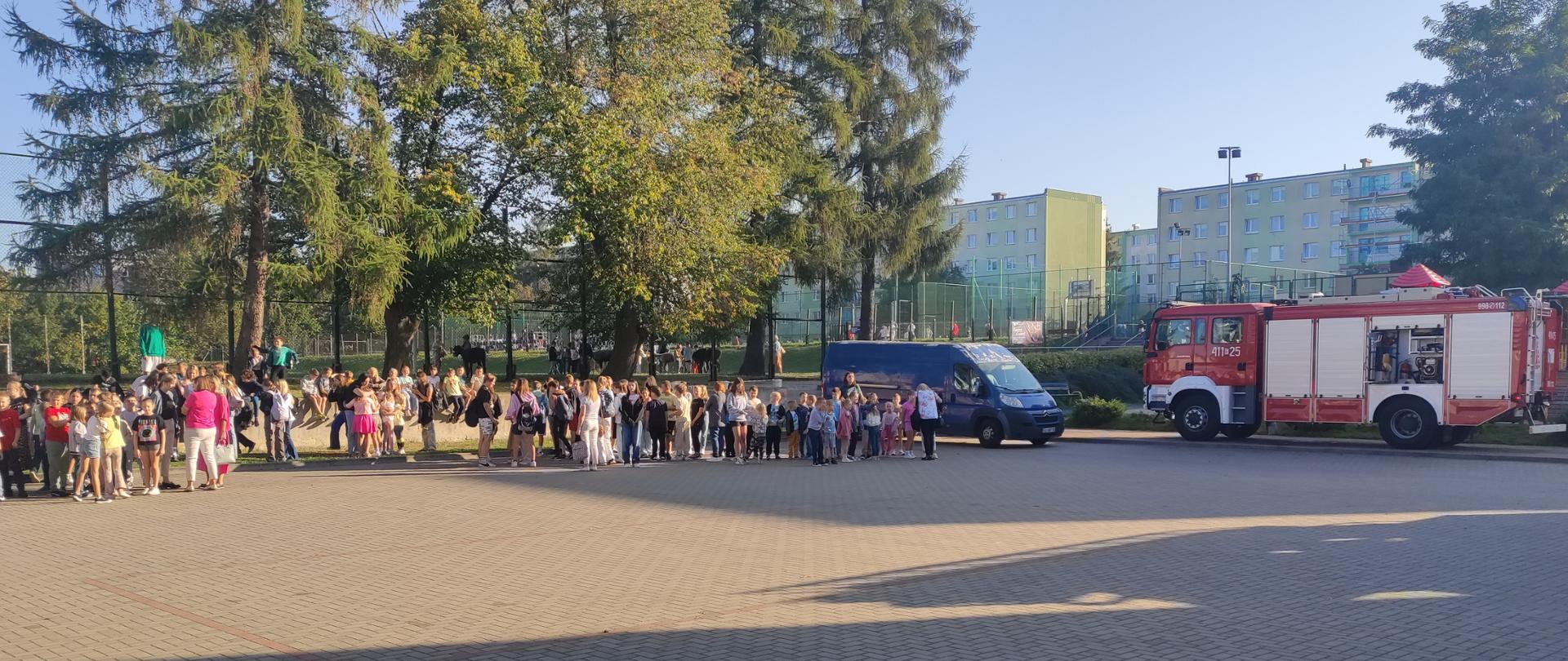 Plac przed szkołą podstawową na placu w punkcie ewakuacyjnym zgromadzone dzieci , dzieci razem ze swoimi wychowawcami w szeregach klasami, dalej po prawej wóż bojowy z jednostki ratowniczo gaśniczej w Brzezinach