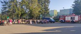 Plac przed szkołą podstawową na placu w punkcie ewakuacyjnym zgromadzone dzieci , dzieci razem ze swoimi wychowawcami w szeregach klasami, dalej po prawej wóż bojowy z jednostki ratowniczo gaśniczej w Brzezinach