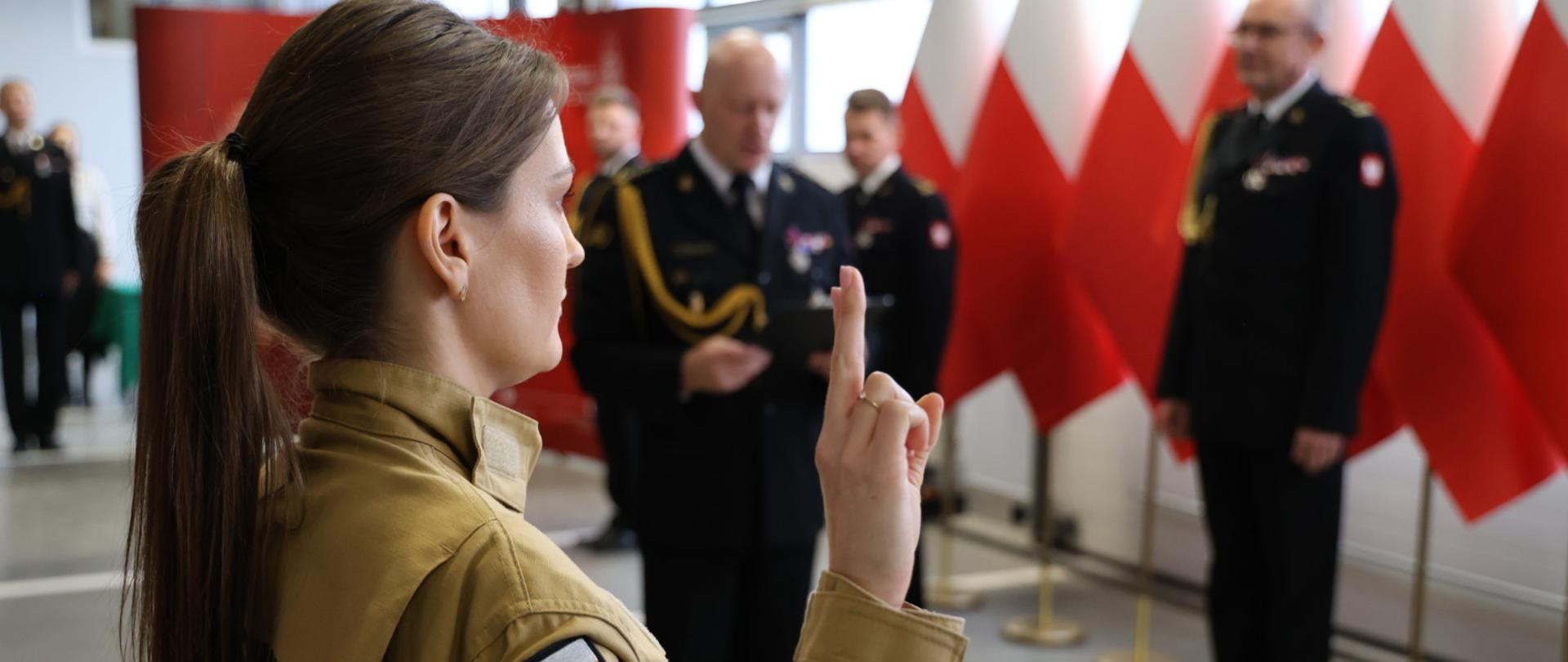 Na pierwszym planie strażaczka w mundurze służbowym, widok z profilu. Trzyma prawą rękę w górze i dwa palce składa do przysięgi. W tle widać strażaków w mundurze galowym, flagi biało-czerwone.