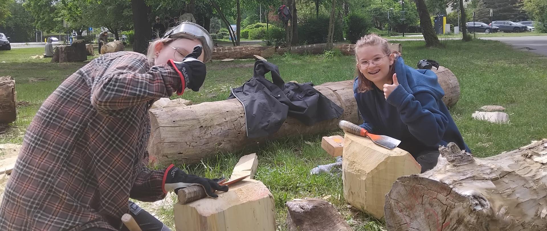 Na zdjęciu widać dwie młode osoby pracujące wspólnie przy rzeźbieniu drewna w plenerze. Obie skupione są na obróbce swoich klocków drewna – jedna klęczy w koszuli w kratę i rękawiczkach, trzymając młotek i dłuto, druga siedzi naprzeciwko w ciemnoniebieskiej bluzie, również pracując dłutem. Wokół nich leżą inne drewniane pnie oraz wióry, a w tle widoczny jest park, droga z zaparkowanymi samochodami i kilka innych osób zajmujących się podobną pracą. Scena emanuje spokojem i koncentracją, typowymi dla warsztatów rzemieślniczych na świeżym powietrzu.