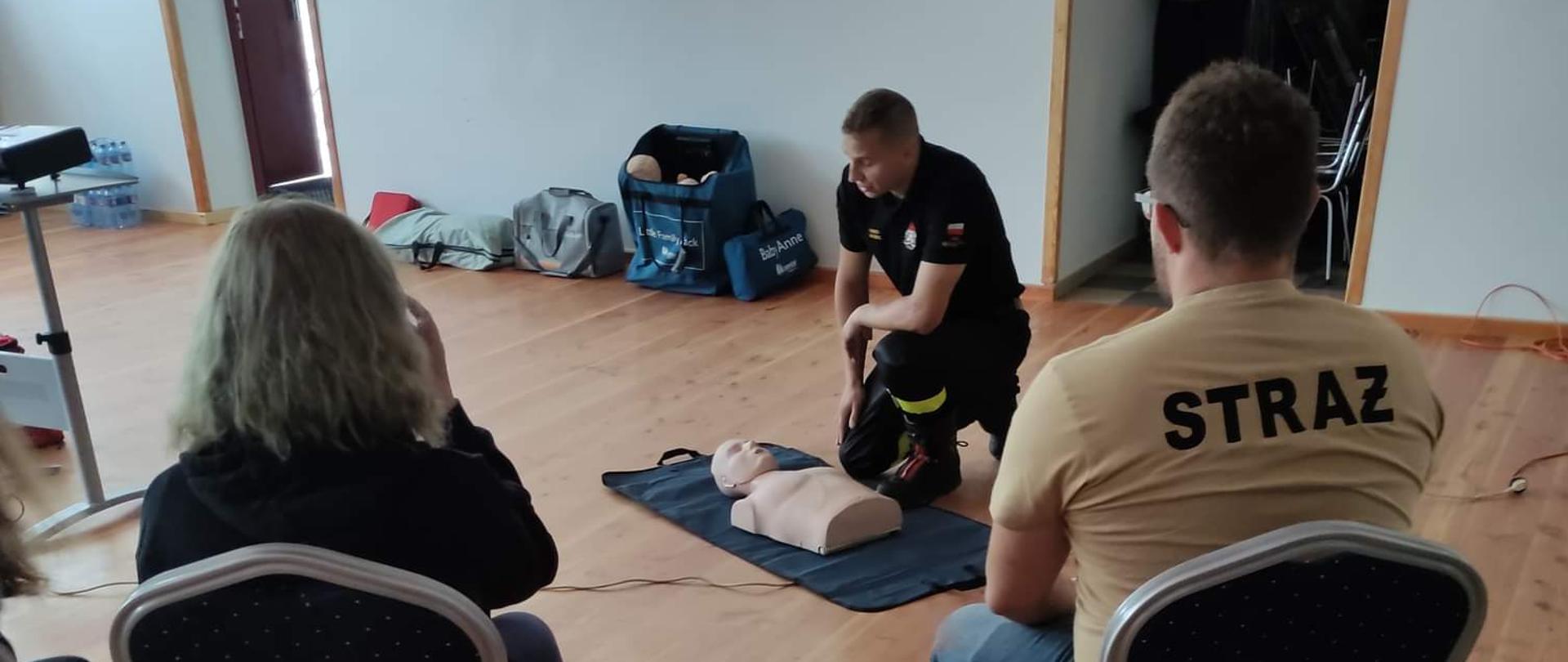 Zdjęcie wykonane na sali wiejskiej w Tłokach. Przedstawia ćwiczących uczestników warsztatów uciskających fantomy. Obok ćwiczących widać strażaka ubranego w mundur koszarowy, tłumaczącego zagadnienia dot. prawidłowego użycia AED.