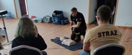 Zdjęcie wykonane na sali wiejskiej w Tłokach. Przedstawia ćwiczących uczestników warsztatów uciskających fantomy. Obok ćwiczących widać strażaka ubranego w mundur koszarowy, tłumaczącego zagadnienia dot. prawidłowego użycia AED.