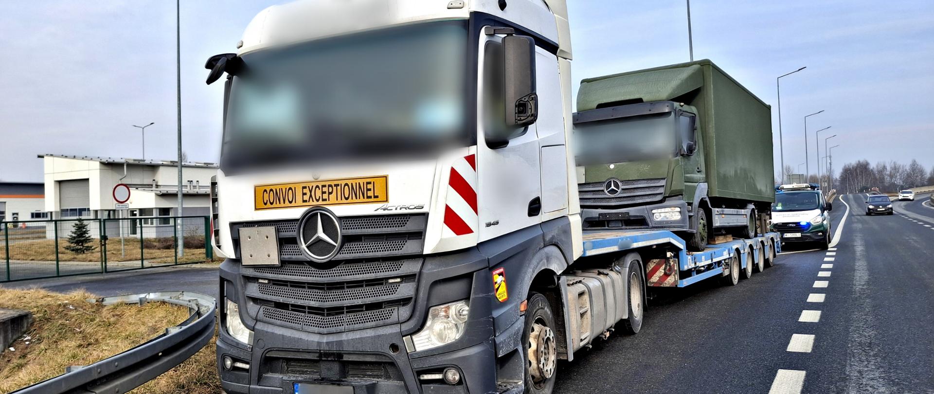Biały ciągnik siodłowy Mercedes-Benz Actros z niskopodwoziową naczepą, zatrzymany na poboczu drogi szybkiego ruchu. Na masce ciężarówki widnieje duża żółta tablica z czarnym napisem "CONVOI EXCEPTIONNEL", wskazująca na transport nienormatywny. Na naczepie przewożona jest mniejsza, zielona ciężarówka w kolorystyce wojskowej. W tle, przed ciężarówką, stoi radiowóz Inspekcji Transportu Drogowego (ITD) z włączonymi sygnałami świetlnymi.