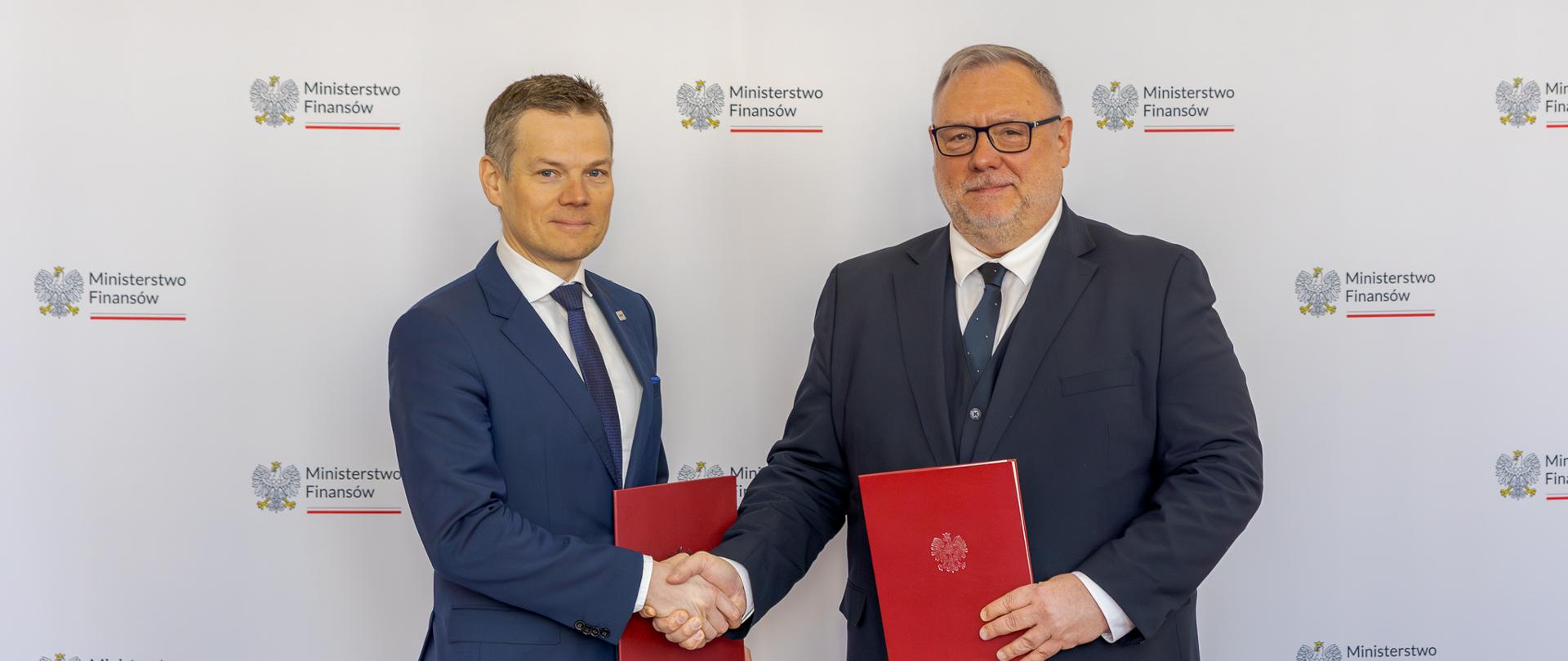 Dwaj mężczyźni podają sobie dłonie, trzymając czerwone teczki z dokumentami, na tle ścianki z logo Ministerstwa Finansów.