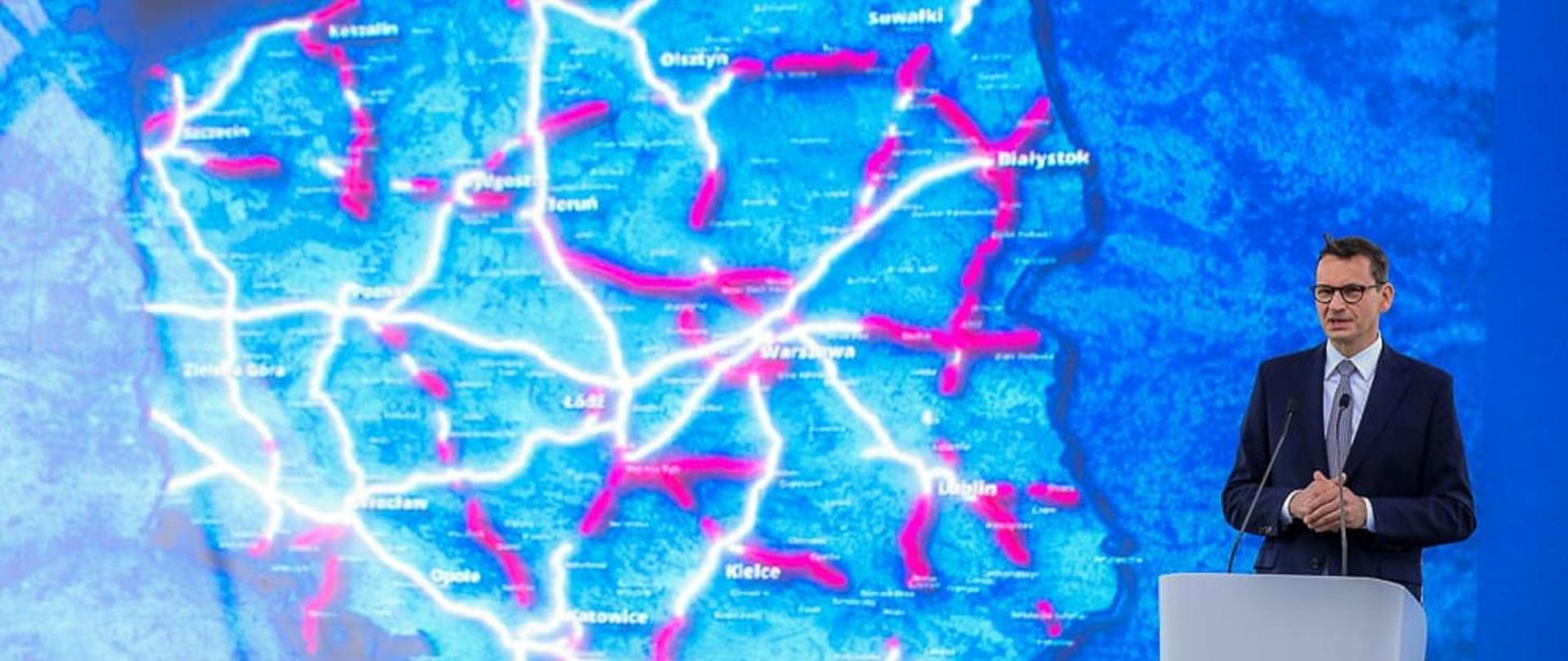 Na pierwszym planie zdjęcia znajduje się elegancko ubrany mężczyzna. Mężczyzna ten stoi za mównicą. W tle znajduje się ekran multimedialny, który przedstawia mapę Polski oraz szlaki drogowe, które zostały zrealizowane oraz te, które są w trakcie realizacji. 