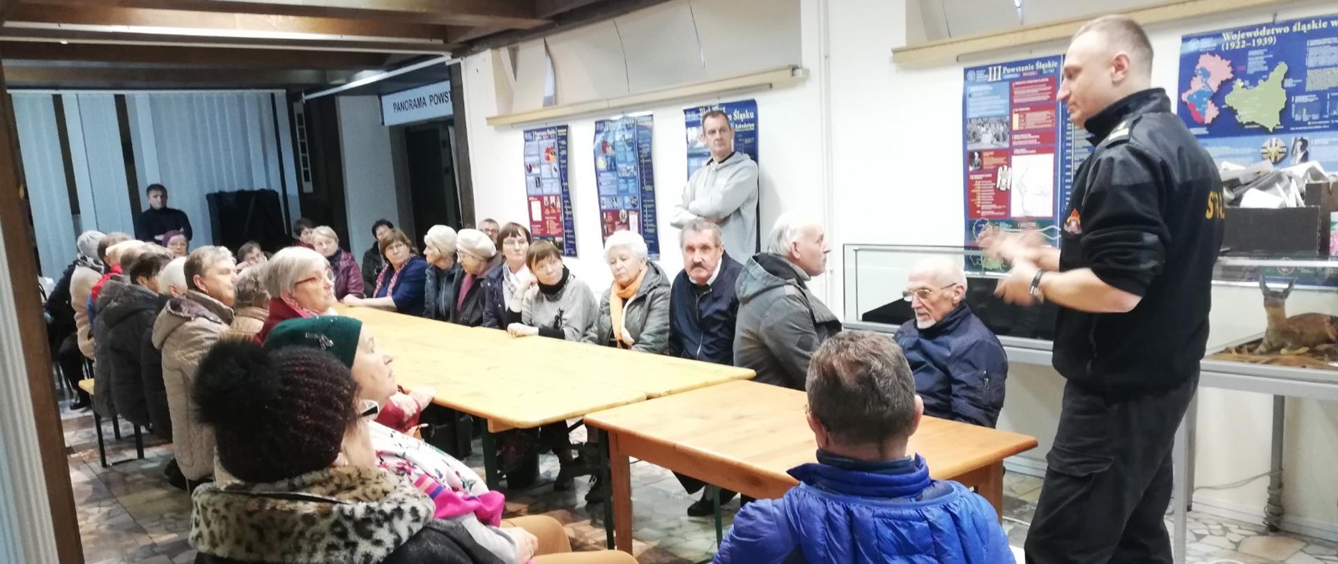 Na zdjęciu znajdują się osoby siedzące przy stołach oraz strażak w ubraniu sztabowym