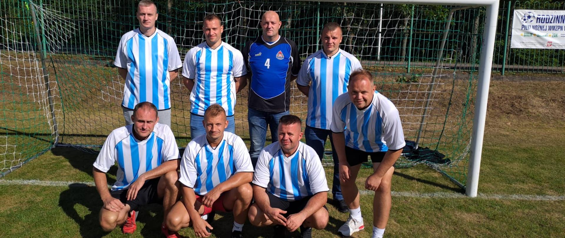 Ośmiu zawodników w biało niebieskich strojach pozują do zdjęcia na tle bramki piłkarskiej. Zielona murawa boiska.