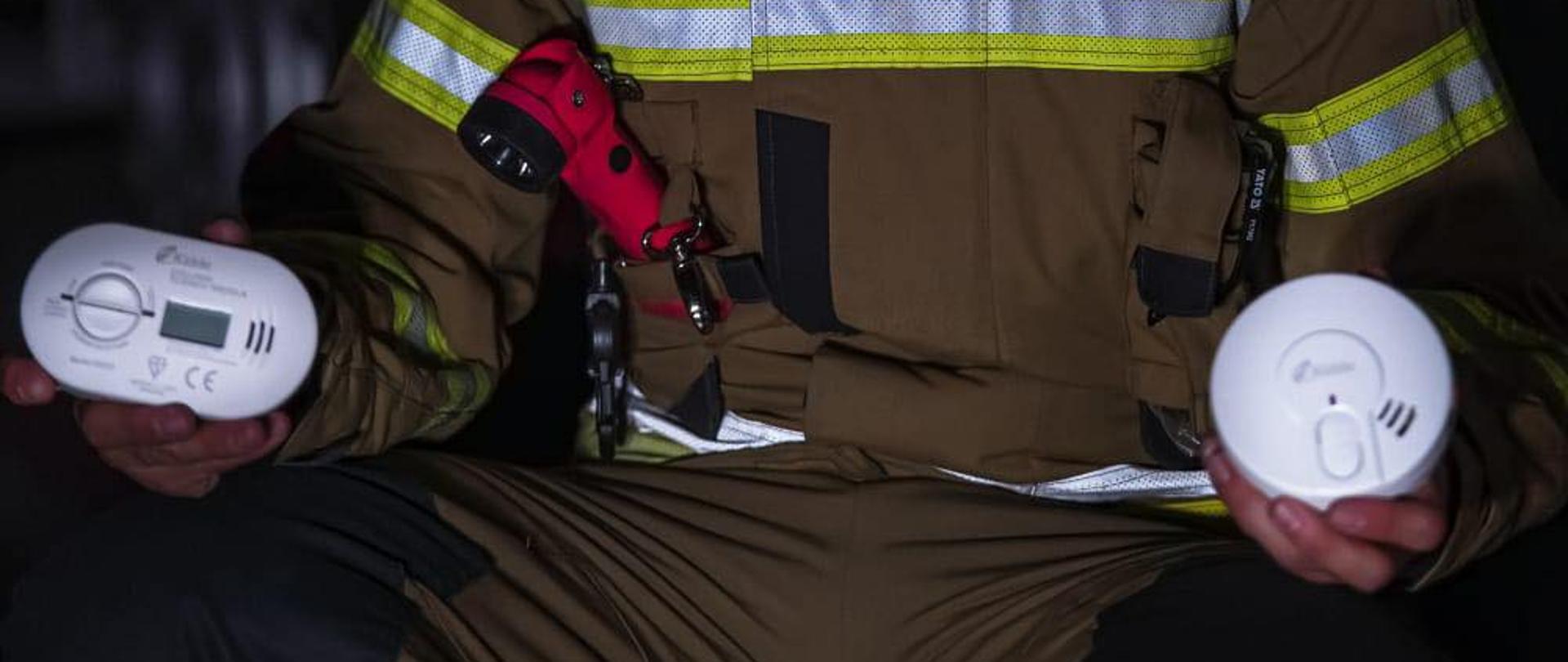 Zdjęcie przedstawia strażaka, który trzyma w ręku dwie czujki (dymu i czadu). 