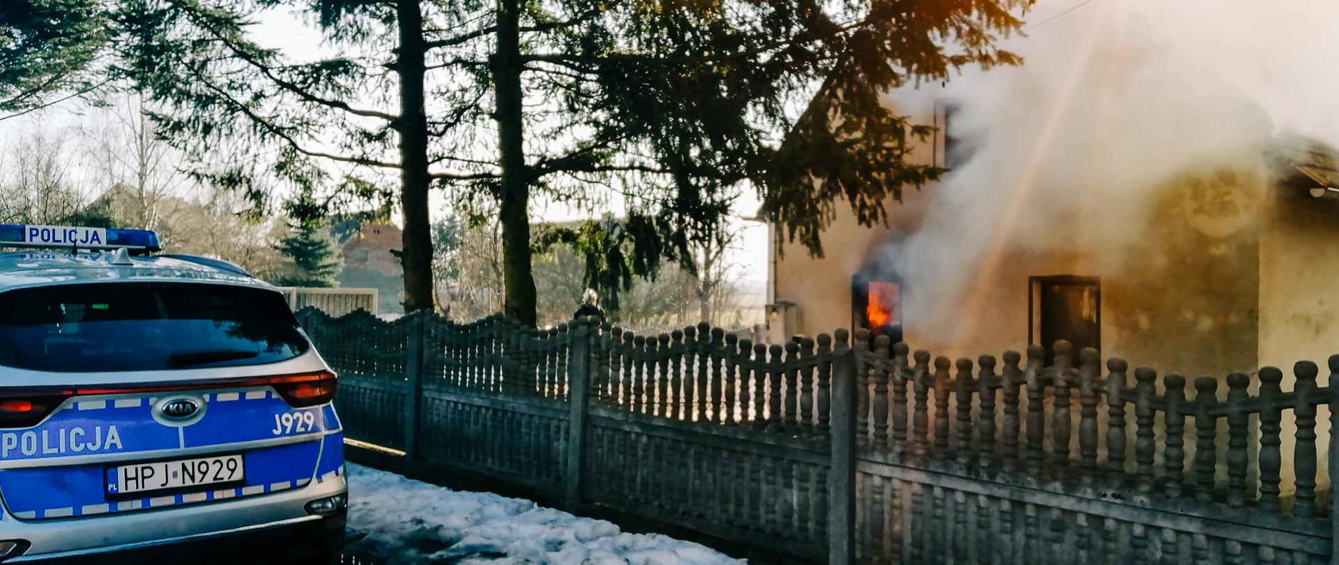 Zdjęcie przedstawia pożar budynku jednorodzinnego. Budynek z widocznym ogniem w jednym z okien, zadymieniem znajduje się po prawej stronie kadru. Centralnie na zdjęciu widoczne dwa świerki przy płocie betonowym - prefabrykowanym. Po lewej stronie widoczny tył radiowozu policji stojący na drodze.