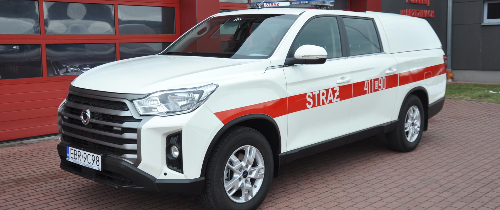 Biały samochód SsangYong na tle budynku komendy powiatowej psp w Brzezinach. Widok na lewy bok.