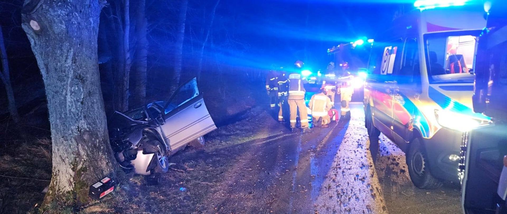 Noc. Niebieskie światła służb ratowniczych. Z lewej strony, częściowo w rowie leży auto osobowe które uderzył w drzewo. Na drodze strazacy i samochody.