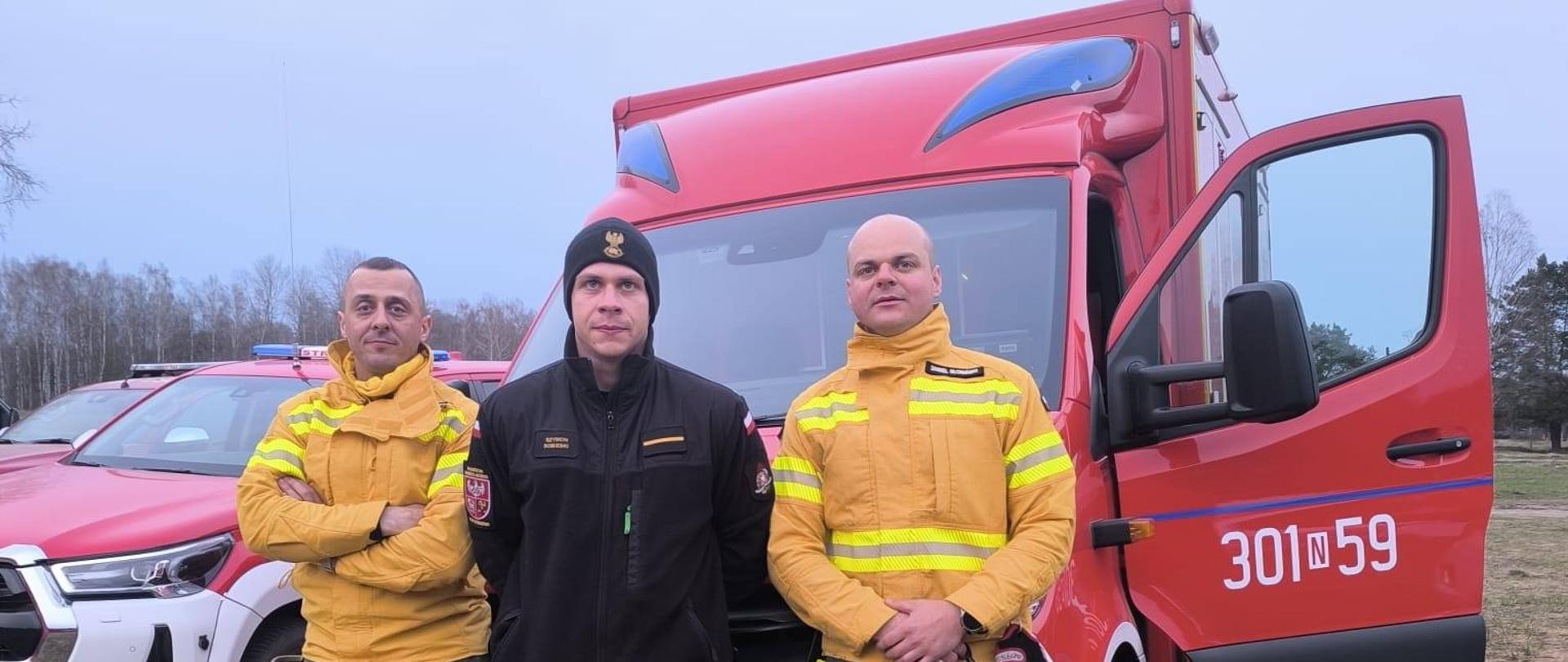 Trzech strażaków na zdjęciu, opierają się o czerwony samochód karetkę strażacką.