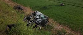 Na zdjęciu widoczny jest uszkodzony pojazd marki BMW. Auto ze zgniecionym przodem znajduje się w połowie przydrożnej skarpy. W tle, w polu ze wschodzącym zboże, w odległości 20 m od pojazdu leży wyrwany silnik.