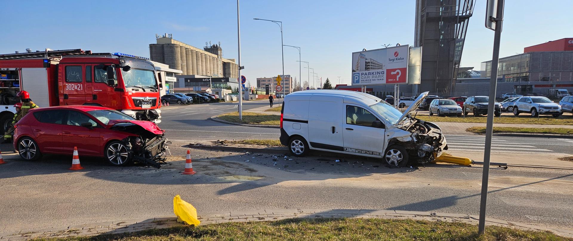 Zdjęcie przedstawia miejsce wypadku: dwa zniszczone auta stojące jeden za drugim. Z lewej strony, za autami stoi samochód pożarniczy i widać dwóch ratowników.