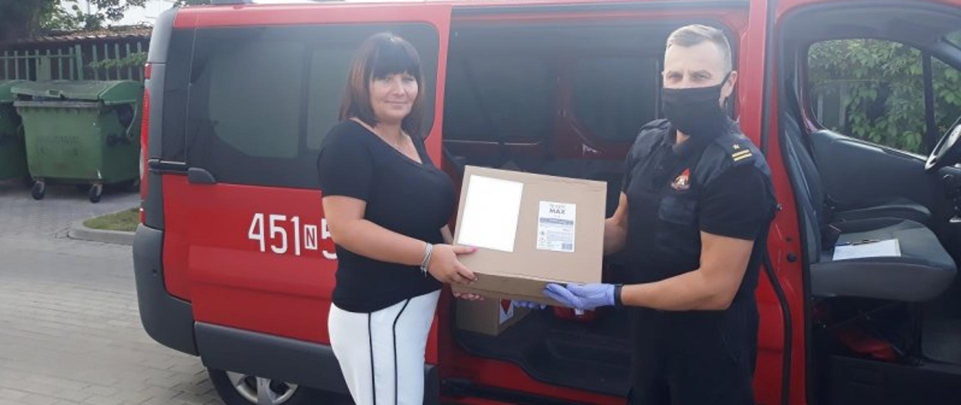 Gołdapscy strażacy przekazali środki do dezynfekcji oraz maseczki ochronne – na zdjęciu strażak przekazuje pudełko z maseczkami ochronnymi na tle samochodu strażackiego. Na drugim planie okoliczne budynki.