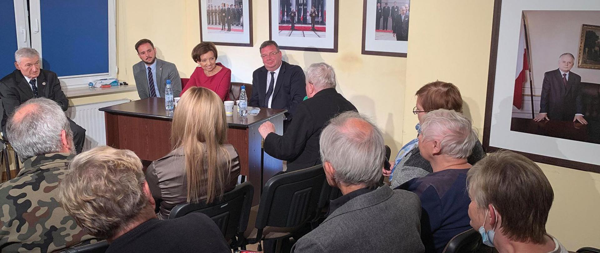 Pomieszczenie. Za stolikiem trzy osoby, wśród nich minister Maląg. Obok na krzesłach siedzą, odwróceniu twarzami uczestnicy spotkania. Na ścianach zdjęcia. Wśród nich zdjęcie prezydenta L. Kaczyńskiego. 