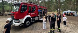 W lesie stoi duży czerwony wóz strażacki. Obok samochodu zebrała się grupa dzieci. Dwóch strażaków w czarnych spodniach z odblaskowymi pasami rozmawia z nimi i pokazuje wyposażenie wozu.
W tle widać niebiesko-białą plandekę rozpiętą nad stołem.