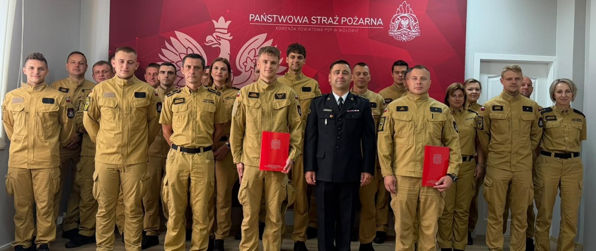 Zdjęcie grupowe funkcjonariuszy Państwowej Straży Pożarnej w Komendzie Powiatowej w Wołowie. Grupa stoi przodem na tle czerwonej ścianki z godłem Polski, logiem PSP i napisem państwowa straż pożarna – komenda powiatowa psp w wołowie. Większość osób nosi piaskowe mundury służbowe, a dwie osoby z przodu trzymają czerwone teczki. Pośrodku stoi oficer w ciemnym mundurze wyjściowym. W grupie są zarówno mężczyźni, jak i kobiety.