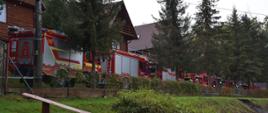 Zdjęcie przedstawia samochody ratowniczo-gaśnicze oraz ratowników podczas ćwiczeń. W tle zwarta zabudowa drewnianych budynków w Ośrodku Wypoczynkowym.
