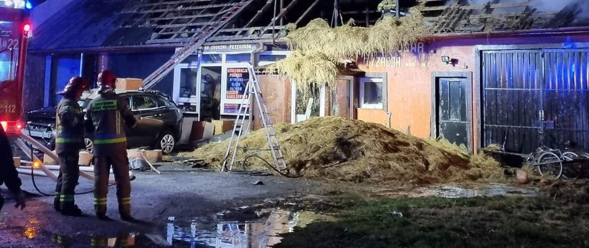 Spalone poddasze warsztatu samochodowego. Przed obiektem znajdują resztki usuniętego z budynku siana. Strażak z linią gaśniczą na stropie dogasza pożar. 