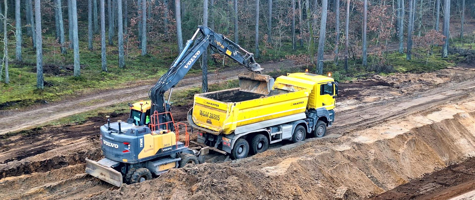Na zdjęciu widać koparkę, która ładuje ziemię na ciężarówkę. Cała scena ma miejsce na placu budowy w lesie, gdzie w tle widać wysokie drzewa. Koparka znajduje się w wykopie, a ciężarówka stoi na drodze obok wykopu.