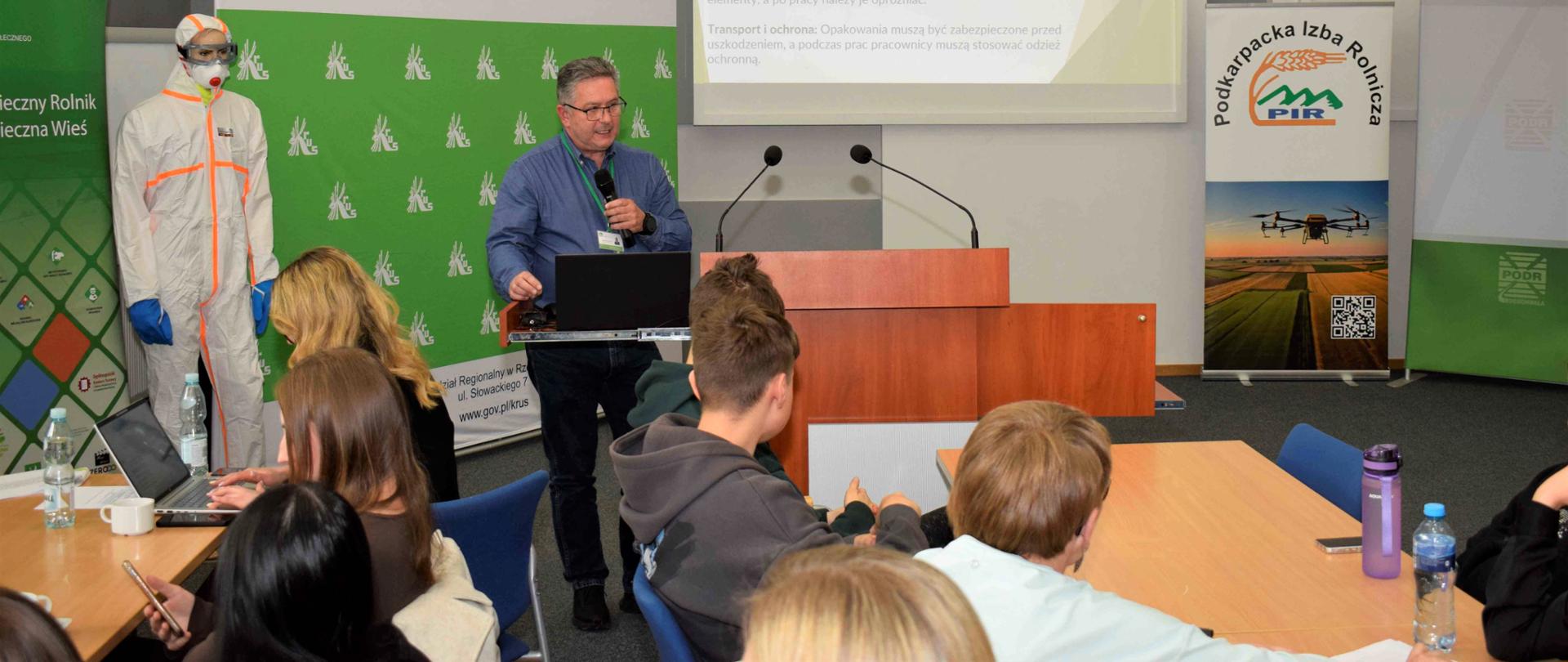 Na sali konferencyjnej przy pulpicie stoi mężczyzna z mikrofonem. Za nim znajdują się roll-upy Kasy oraz ekran z wyświetloną prezentacją multimedialną dotycząca BHP w rolnictwie.