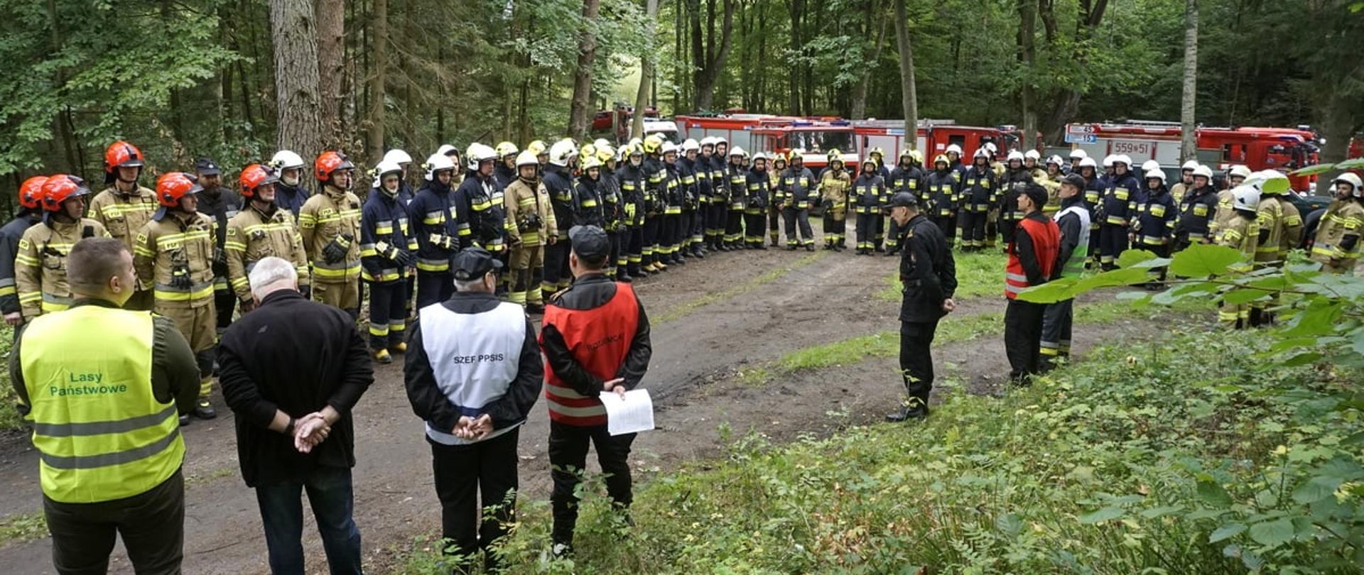 Zdjęcie przedstawia zbiórkę przed ćwiczeniami "Lasy 2021". Zbiórka jest zorganizowana w lesie. Przemówienia udziela Komendant Powiatowy państwowej Straży pożarnej w Świdwinie.