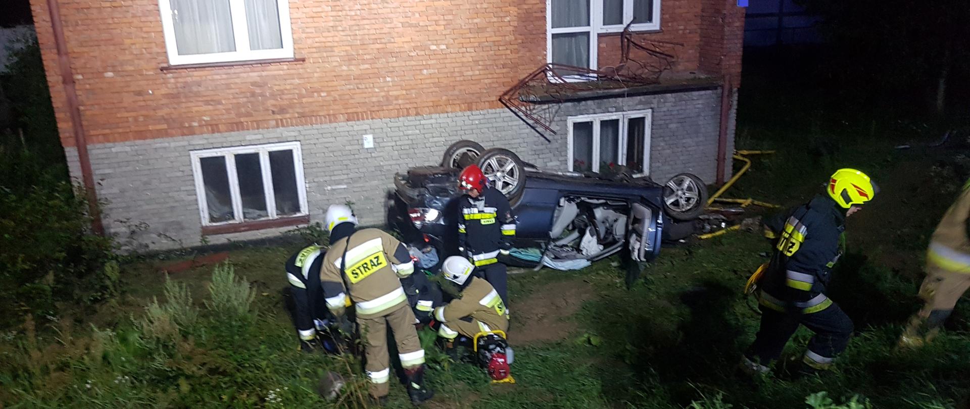 Na zdjęciu widać dom jednorodzinny przed którym do góry kołami leży samochód osobowy. Przed budynkiem obok wraku samochodu 4 strażaków udziela pierwszej pomocy osobie poszkodowanej. Zdjęcie wykonane ze wzniesienia przed prywatna posesją w nocy. Teren akcji jest oświetlony. 