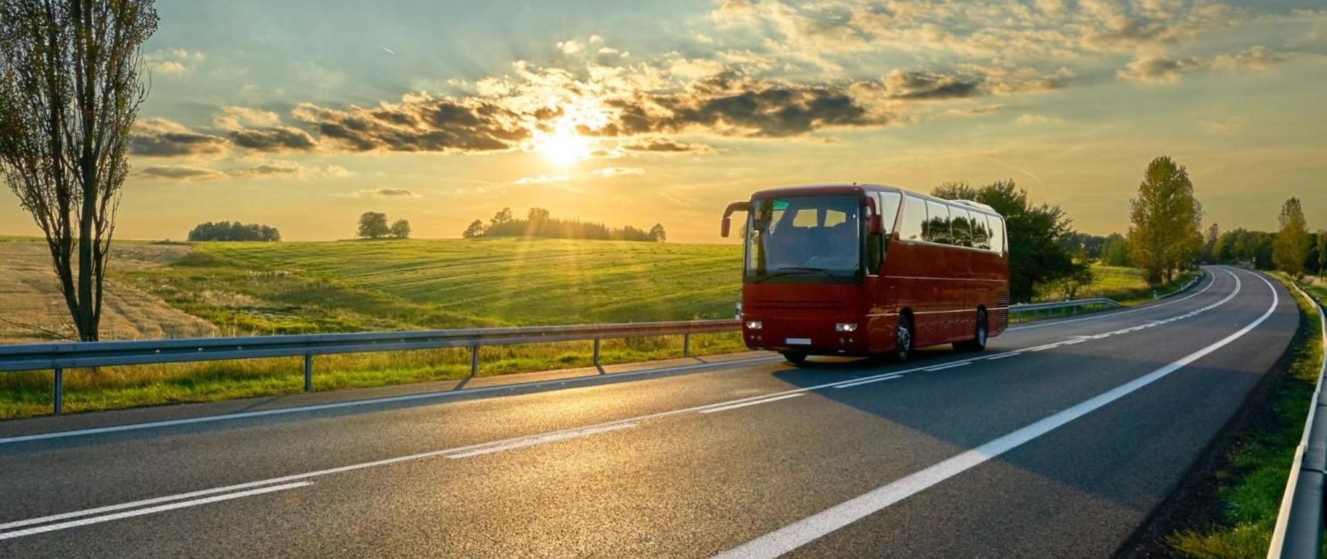 droga asfaltowa, na niej czerwony autobus, w tle zachodzące za chmury słonce