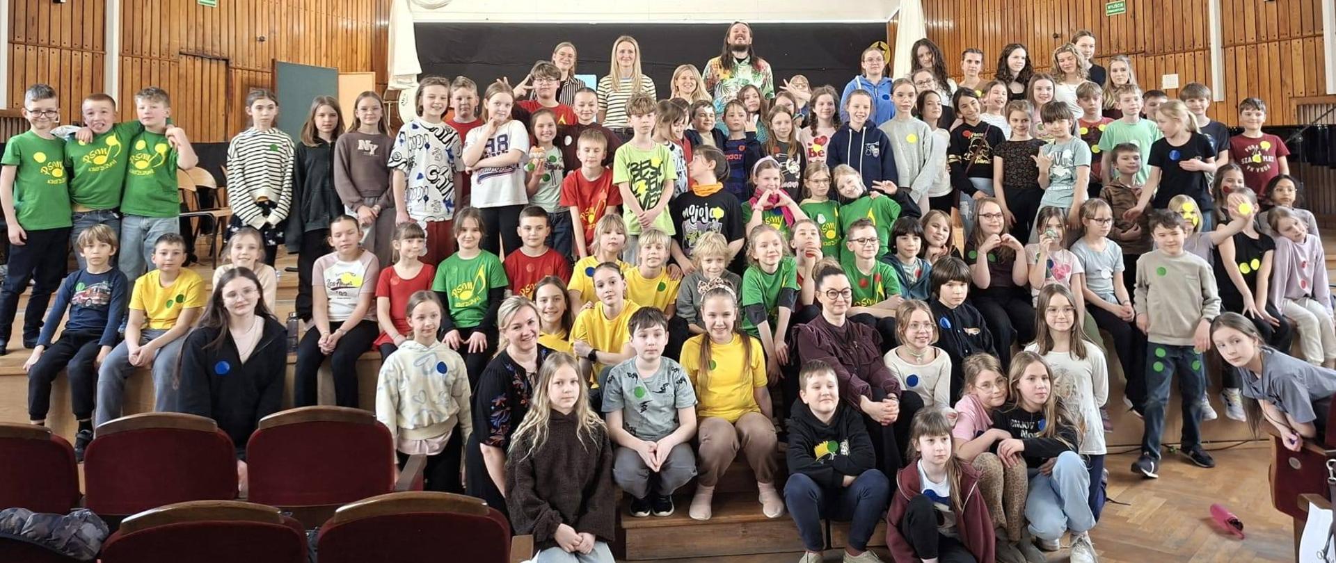 Duża grupa dzieci i kilkoro dorosłych pozuje do wspólnego zdjęcia na scenie w auli z drewnianymi panelami na ścianach. Wiele dzieci ma na sobie kolorowe koszulki (zielone, żółte, czerwone) z okrągłymi naklejkami.