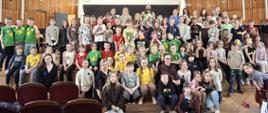 Duża grupa dzieci i kilkoro dorosłych pozuje do wspólnego zdjęcia na scenie w auli z drewnianymi panelami na ścianach. Wiele dzieci ma na sobie kolorowe koszulki (zielone, żółte, czerwone) z okrągłymi naklejkami.