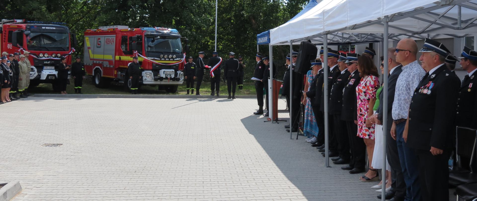 w oddali 2 samochody ratowniczo-gaśnicze po prawej strażacy, druhowie, samorządowcy i inni ludzie stoją pod namiotami bankietowymi