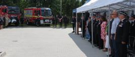 w oddali 2 samochody ratowniczo-gaśnicze po prawej strażacy, druhowie, samorządowcy i inni ludzie stoją pod namiotami bankietowymi