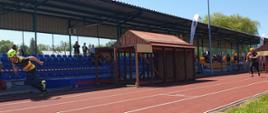 Na zdjęciu widać strażaków w ubraniach sportowych którzy biegną po stadionie z nawierzchnią tartanową po zeskoku z drewnianych domków. W tle trybuny stadionu na których siedzą osoby. 