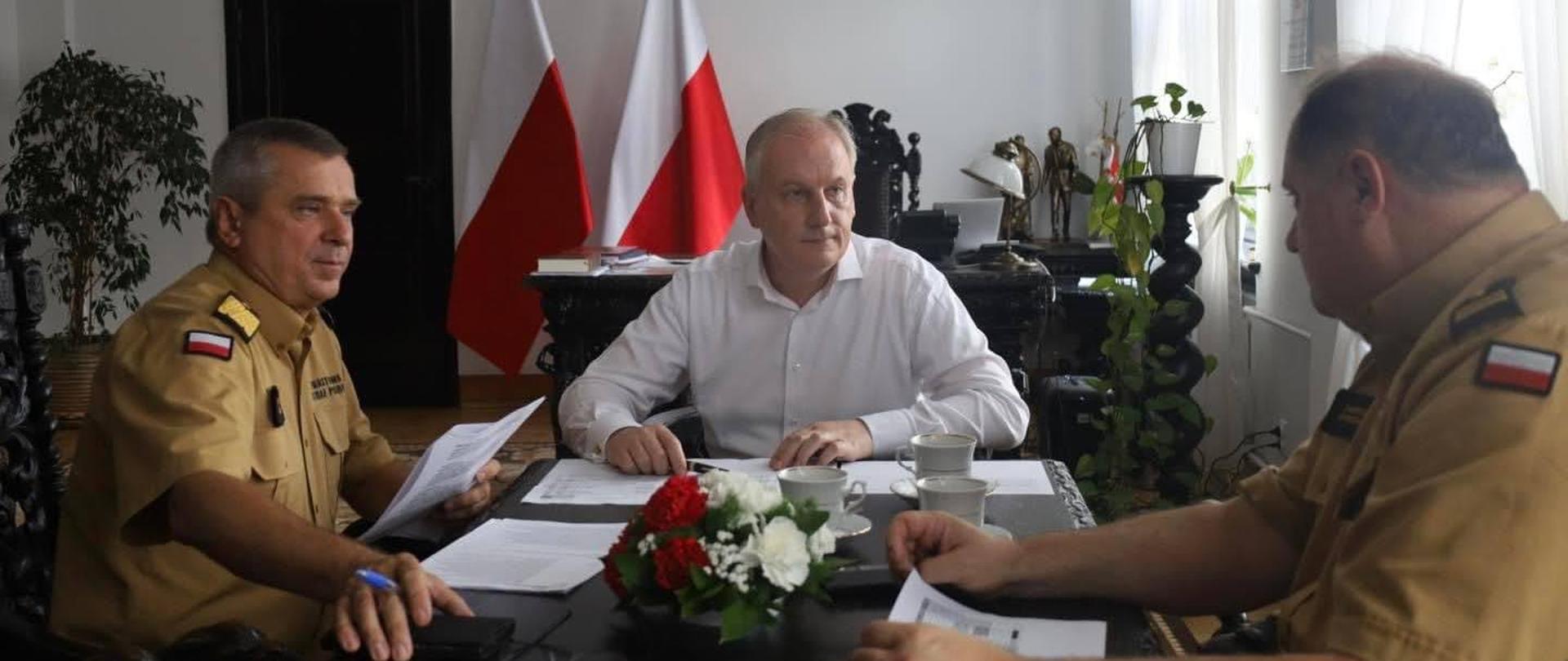 Funkcjonariusze Państwowej Straży Pożarnej w mundurach w kolorze musztardowym siedzą przy stole trzymając karki i rozmawiając z mężczyzną ubranym w białą koszulę na stole leżą kartki papieru oraz ustawione są filiżanki oraz stoi wiązanka kwiatów za nimi stoi biurko oraz ustawione są dwie flagi Polski oraz figurki.