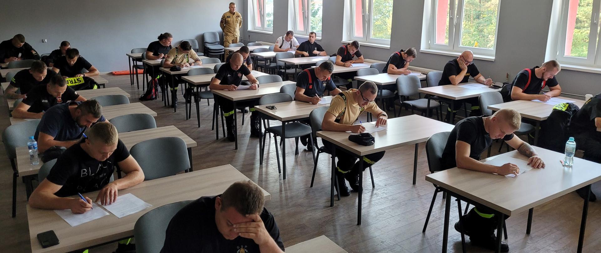 Strażacy siedzący pojedynczo przy stołach na sali wykładowej podczas egzaminu teoretycznego.