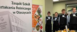 Zdjęcie wykonane z perspektywy wzdłuż długiego stoiska degustacyjnego przykrytego czarnym obrusem. Na stole w równych rzędach ułożono czarne tace oraz koszyki pełne wykwintnych przekąsek: faszerowanych babeczek, kruchych rogalików z wytrawnym nadzieniem, koreczków, sałatek w małych wyporcjowanych pojemniczkach, deski serów oraz puszystych bułeczek. Na stole ustawiono również tabliczki z opisem serwowanych potraw i menu.
W tle, po prawej stronie za stołem, stoi czworo uczniów w galowych strojach kelnerskich – w czarnych kamizelkach, białych koszulach i czarnych muszkach – z dumą prezentujących przygotowane potrawy. 