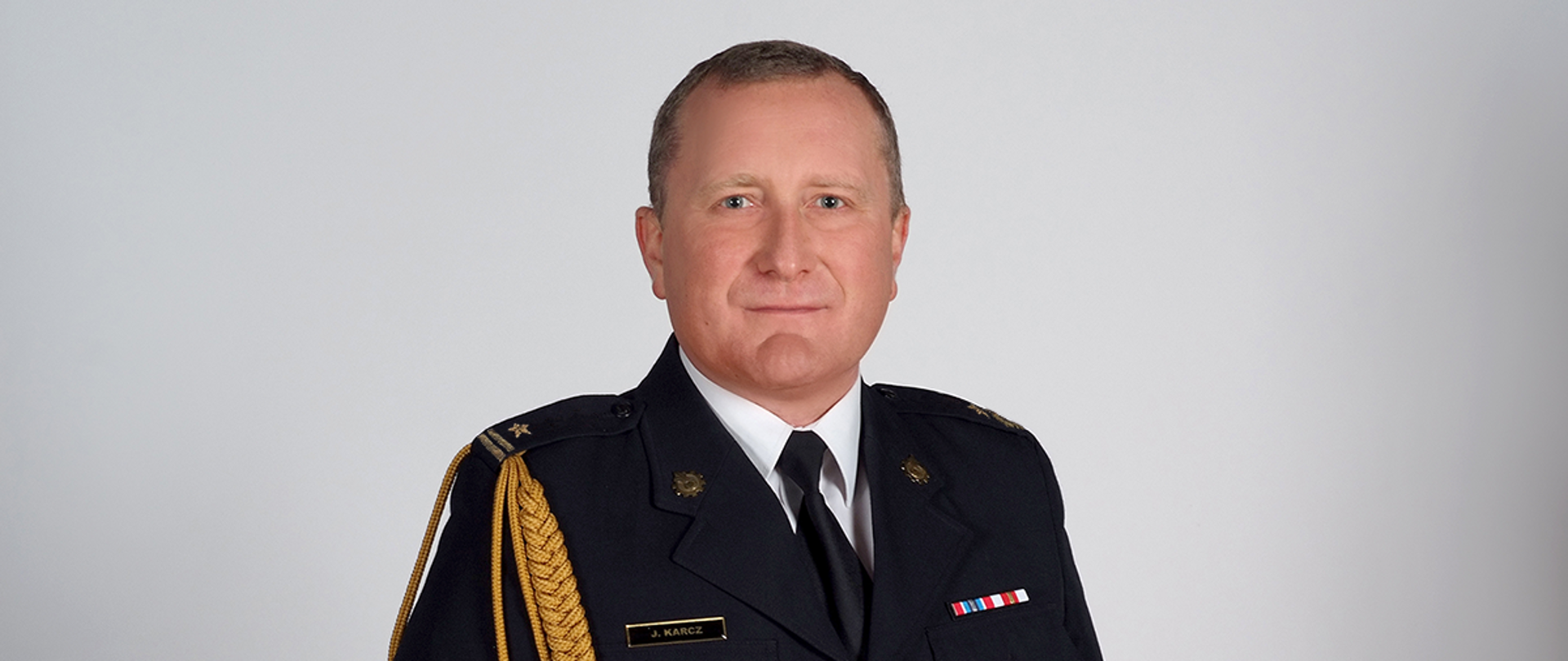 Na zdjęciu młodszy brygadier Jacek Karcz, Komendant Powiatowy Państwowej Straży Pożarnej w Pabianicach. Występuje w umundurowaniu wyjściowym ze sznurem. Na lewej piersi przypięte baretki. Na prawej piersi od góry znajduje się imiennik, absolwentka Szkoły Głównej Służby Pożarniczej oraz absolwentka Szkoły Aspirantów w Poznaniu.